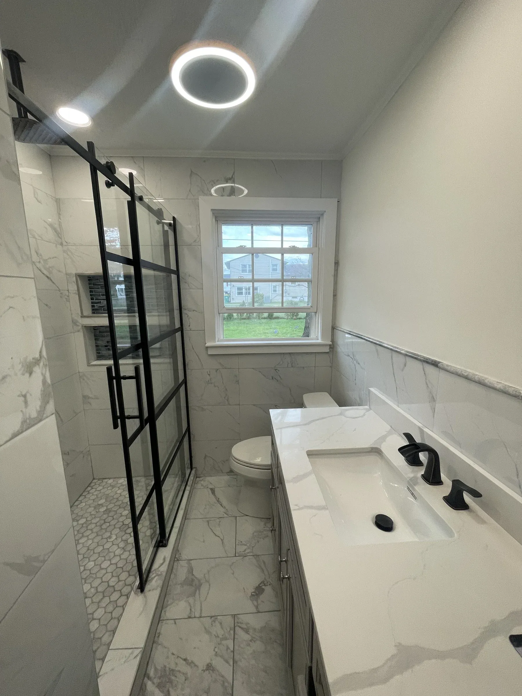 Modern bathroom remodel with marble tile, matte-black fixtures, barn-style glass shower door and circular LED backlit mirror